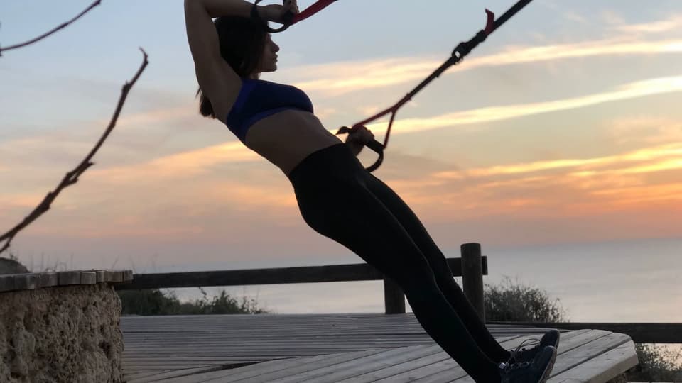 Athlete doing suspension-trainer strength work outdoors at sunset by the sea